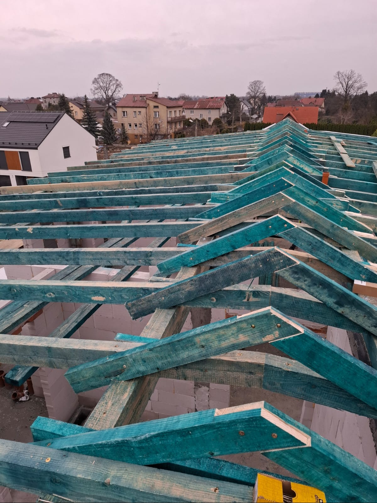 Konstrukcja dachu z impregnowanego drewna na tle zabudowy miejskiej. Widok z góry na szkielet dachu z elementami muru. Niebo pochmurne.