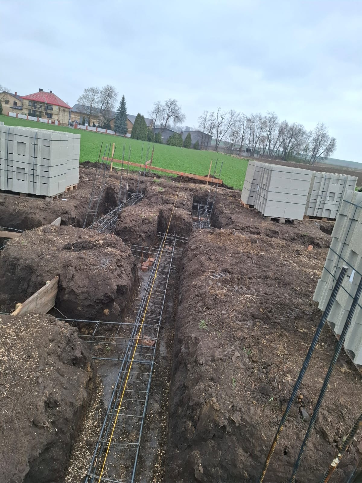 Wykopy pod fundamenty z ułożonym zbrojeniem i bloczkami betonowymi w tle, widok z góry na plac budowy w pochmurny dzień.