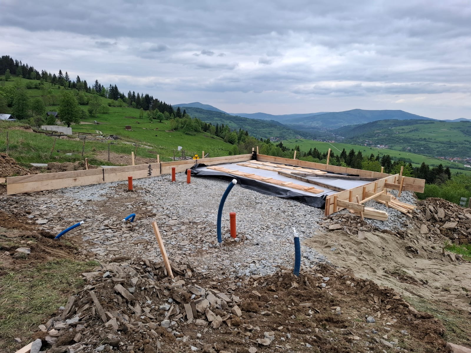 Przygotowanie fundamentów pod dom na zboczu: szalunek z desek, podsypka żwirowa, rury instalacyjne. W tle zielone wzgórza i pochmurne niebo.