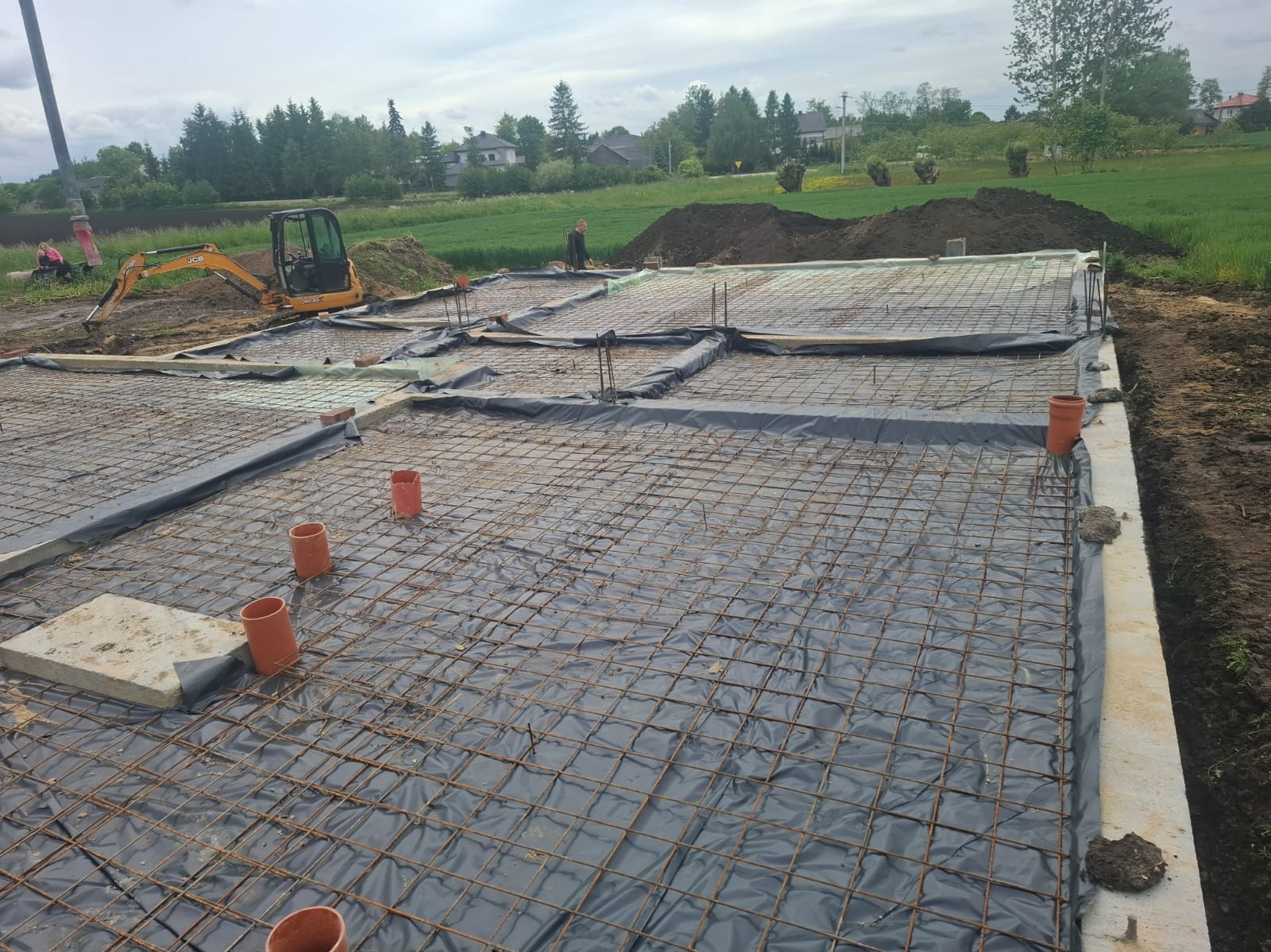 Zbrojenie fundamentów domu jednorodzinnego z widocznym szalunkiem, folią izolacyjną i wystającymi rurami kanalizacyjnymi. W tle koparka i teren budowy.