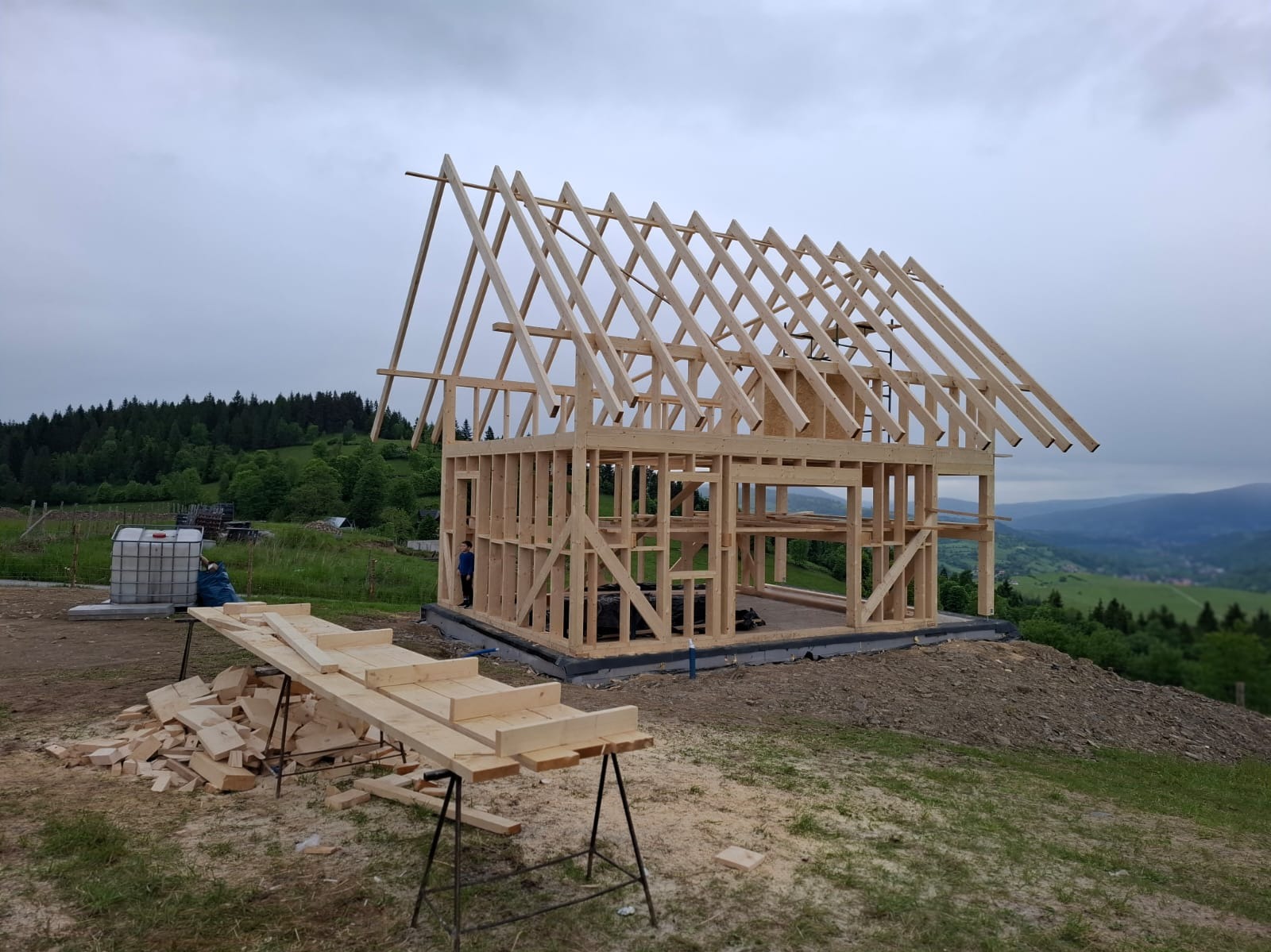 Konstrukcja domu szkieletowego w trakcie budowy na tle górskiego krajobrazu. Widoczne drewniane elementy, rusztowanie i przygotowane deski.