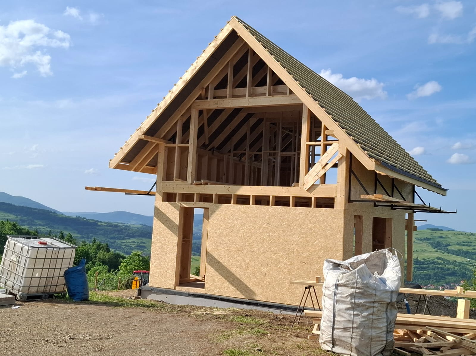 Surowy stan domu szkieletowego z częściowo pokrytym dachem na tle górskiego krajobrazu. Widoczne elementy konstrukcyjne i materiały budowlane.