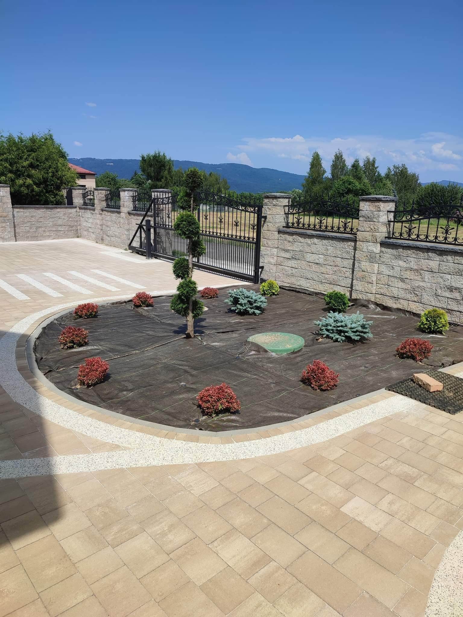 Ogród frontowy z ozdobnymi krzewami i drzewkiem formowanym, wyłożony kostką brukową w odcieniach beżu, z widoczną bramą wjazdową i ogrodzeniem w tle krajobraz górski.