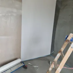 Fragment pokoju w trakcie remontu: gładka, biała tapeta na ścianie kontrastuje z surowym betonem i przygotowaną do tapetowania ścianą; widoczna drabina z plamami farby i rolka tapety na specjalnym...