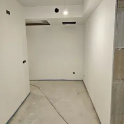 Wnętrze w trakcie prac wykończeniowych: świeżo wyszpachlowane i pomalowane na biało ściany zabezpieczone taśmą malarską przy podłodze, widoczne gniazdka elektryczne i surowy betonowy filar.