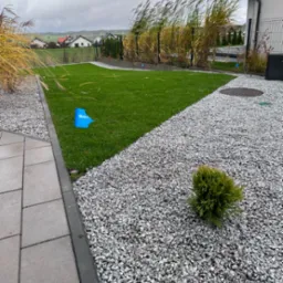 Nowo założony trawnik z rolki, obrzeża z kostki, ścieżka z drobnego grysu, ozdobne trawy wzdłuż ogrodzenia, zieleń na tle pochmurnego nieba.