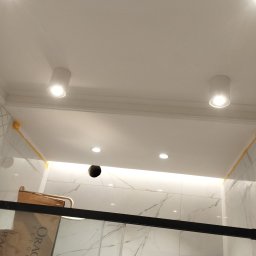 El-pro Łukasz Smoleń - Biała łazienka z marmurowymi płytkami, oświetleniem sufitowym punktowym i listwą LED. Fragment kabiny prysznicowej z czarnym profilem. Widoczne elementy instalacji.