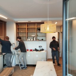 El-pro Łukasz Smoleń - Montaż lampy wiszącej w Krakowie. Dwóch mężczyzn na drabinie, narzędzia na blacie. W tle półki i trzeci mężczyzna. Nowoczesne wnętrze w trakcie wykańczania.