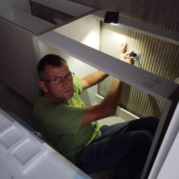 El-pro Łukasz Smoleń - Elektryk w trakcie pracy, montuje gniazdko w szafce podświetlonej lampką. Widoczna ściana z tapetą w tle.