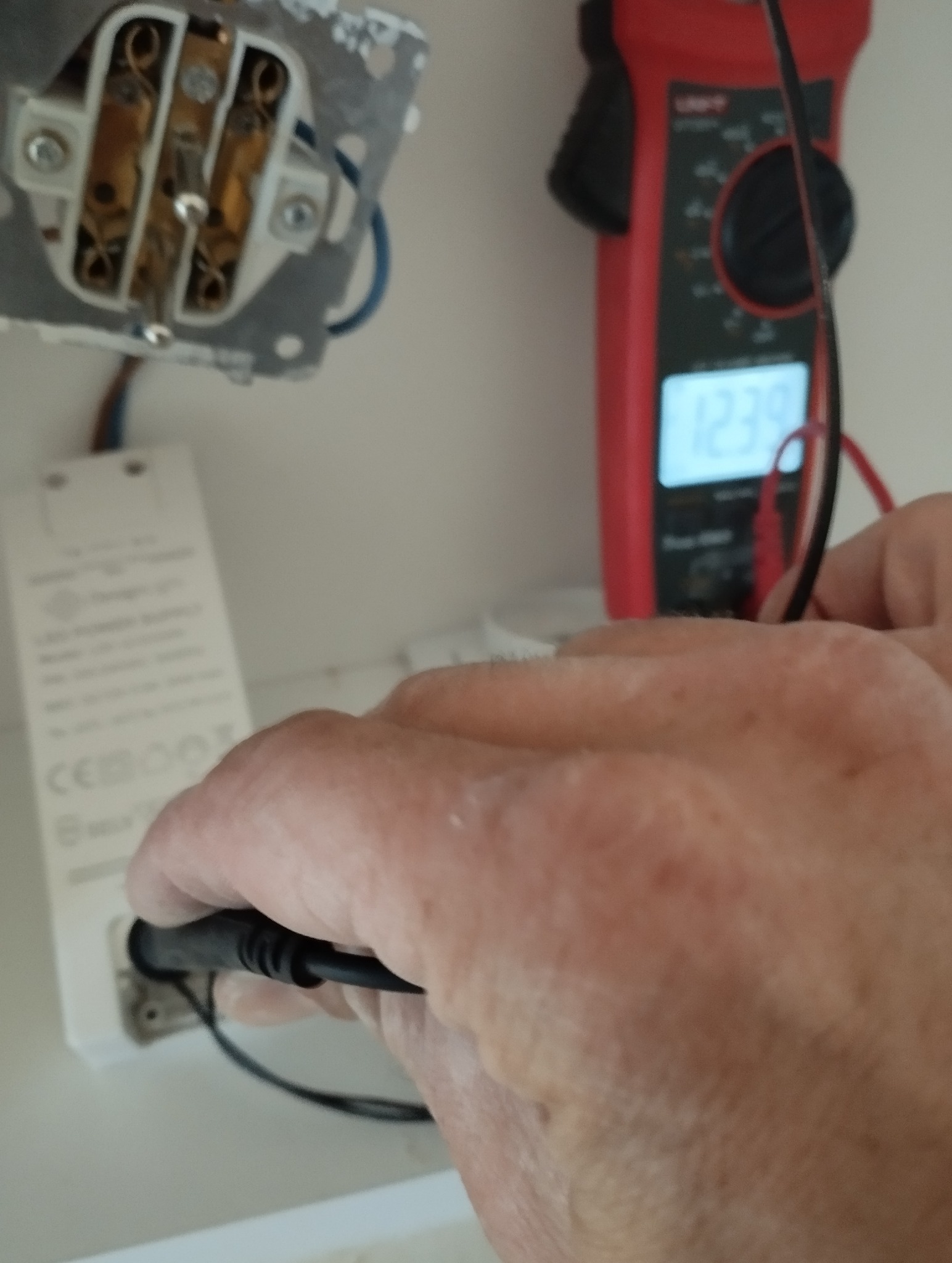 Pomiar napięcia multimetrem na listwie LED, widoczna puszka elektryczna. Przewody podłączone, wyświetlacz multimetru wskazuje wartość.