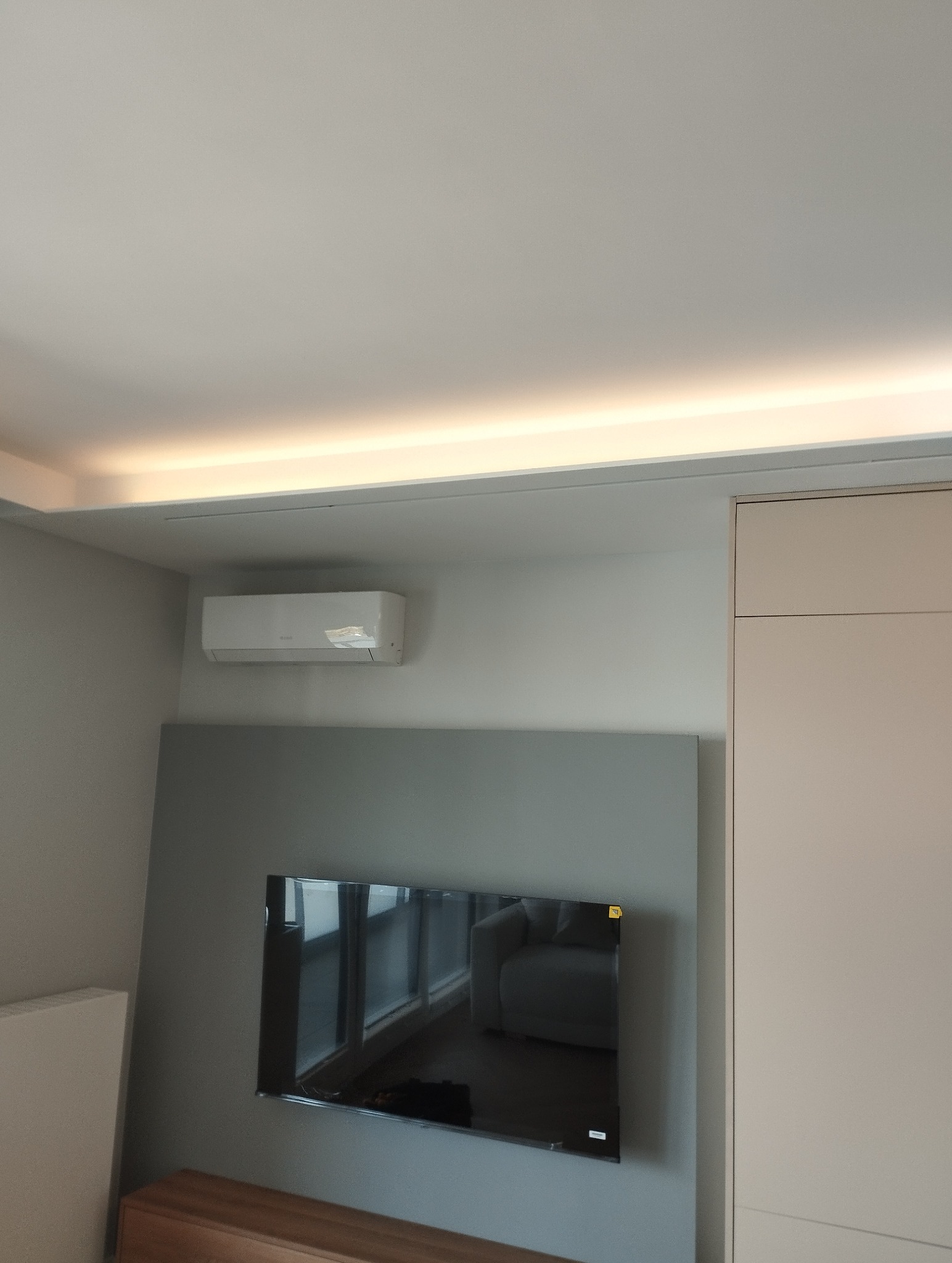 Minimalistyczne wnętrze: podświetlany sufit, klimatyzator na ścianie i telewizor z odbiciem sofy. Nowoczesny design w jasnych barwach.