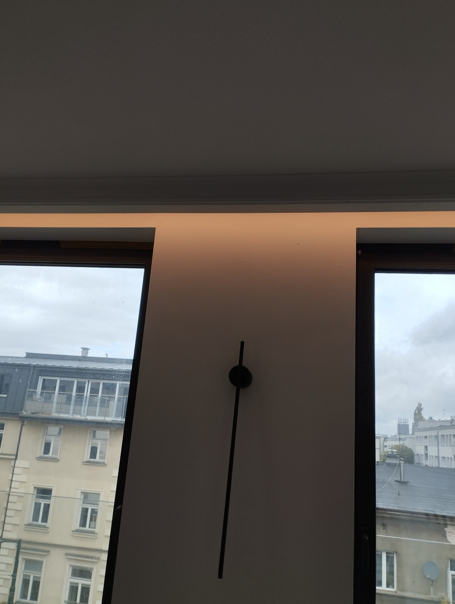 Minimalistyczna lampa ścienna LED, oświetlająca ścianę w ciepłym odcieniu, widok przez okno na budynki w tle. Nowoczesne wnętrze.