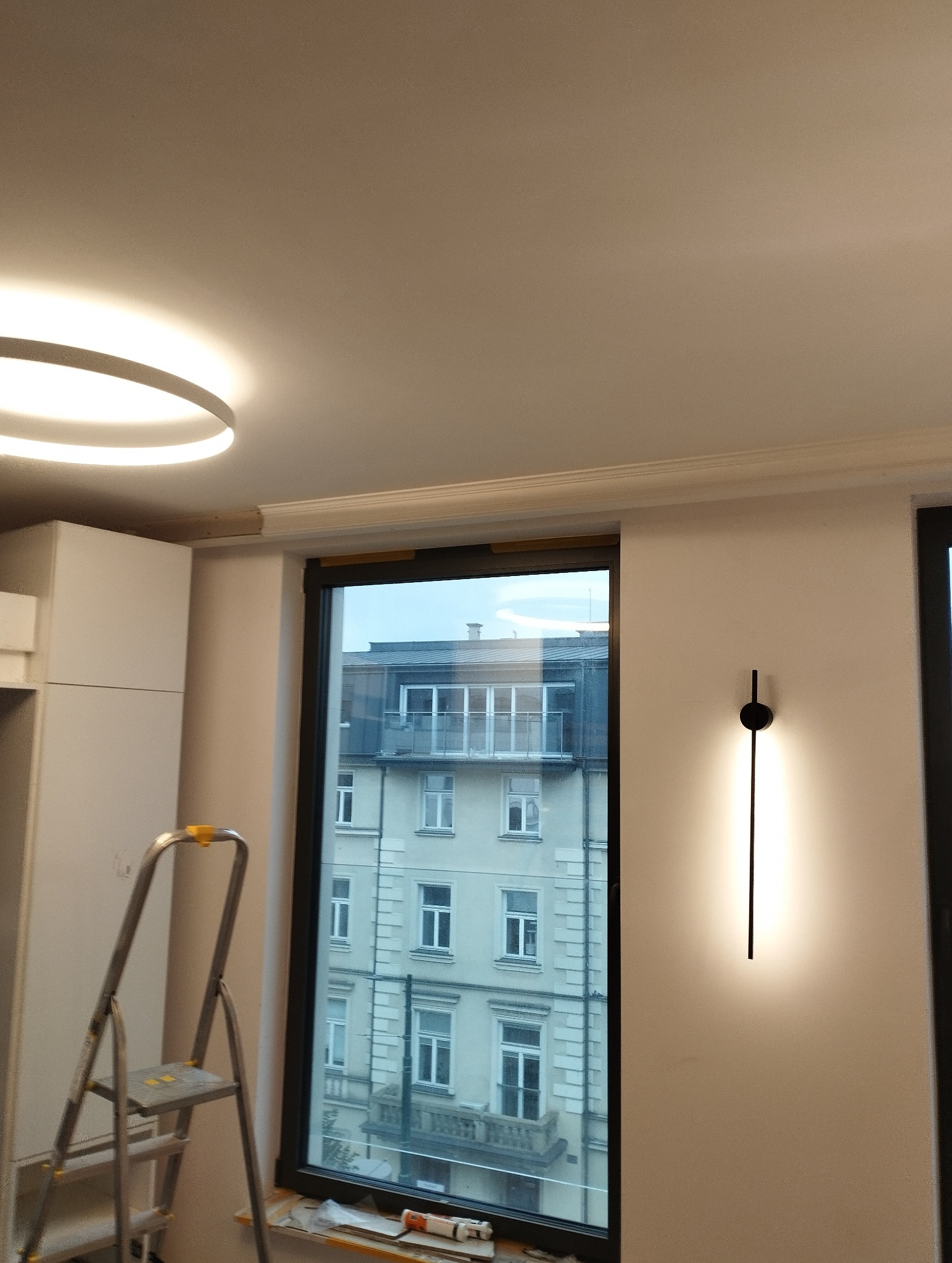 Nowoczesna lampa ścienna zamontowana obok okna z widokiem na budynek. Widoczna drabina i fragment sufitu z oświetleniem. Minimalistyczny design wnętrza w trakcie wykończenia.