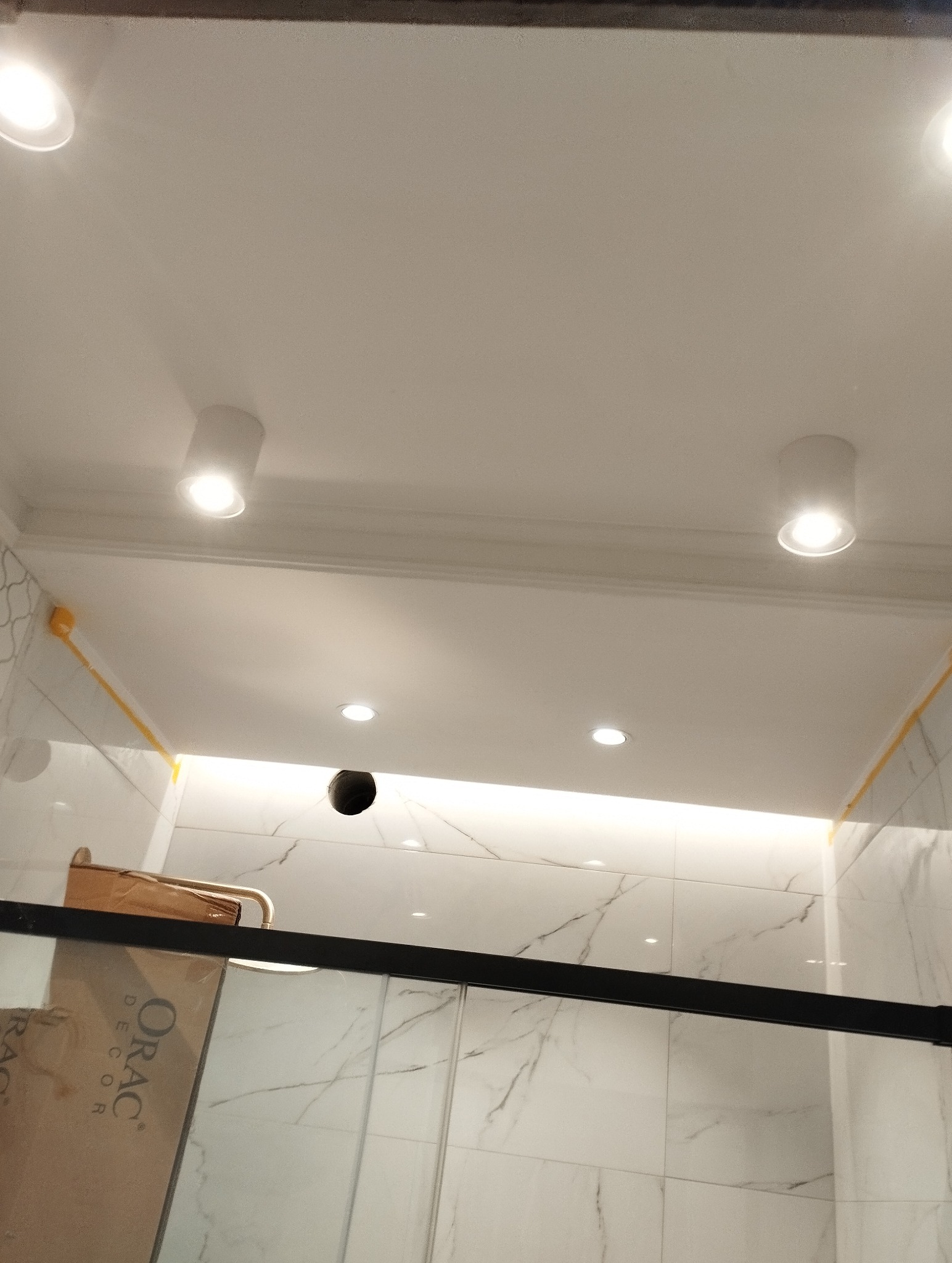 Biała łazienka z marmurowymi płytkami, oświetleniem sufitowym punktowym i listwą LED. Fragment kabiny prysznicowej z czarnym profilem. Widoczne elementy instalacji.