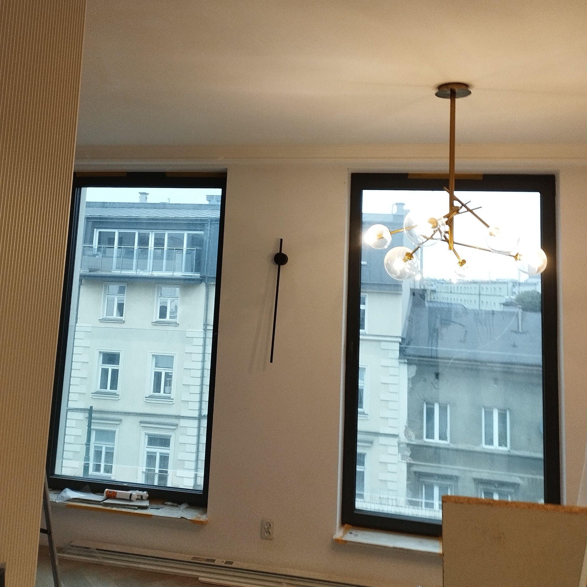 Nowoczesny żyrandol kulisty zamontowany w pokoju z dwoma oknami z widokiem na budynek. Na ścianie minimalistyczny zegar bez cyfr. Widoczne ślady prac wykończeniowych.