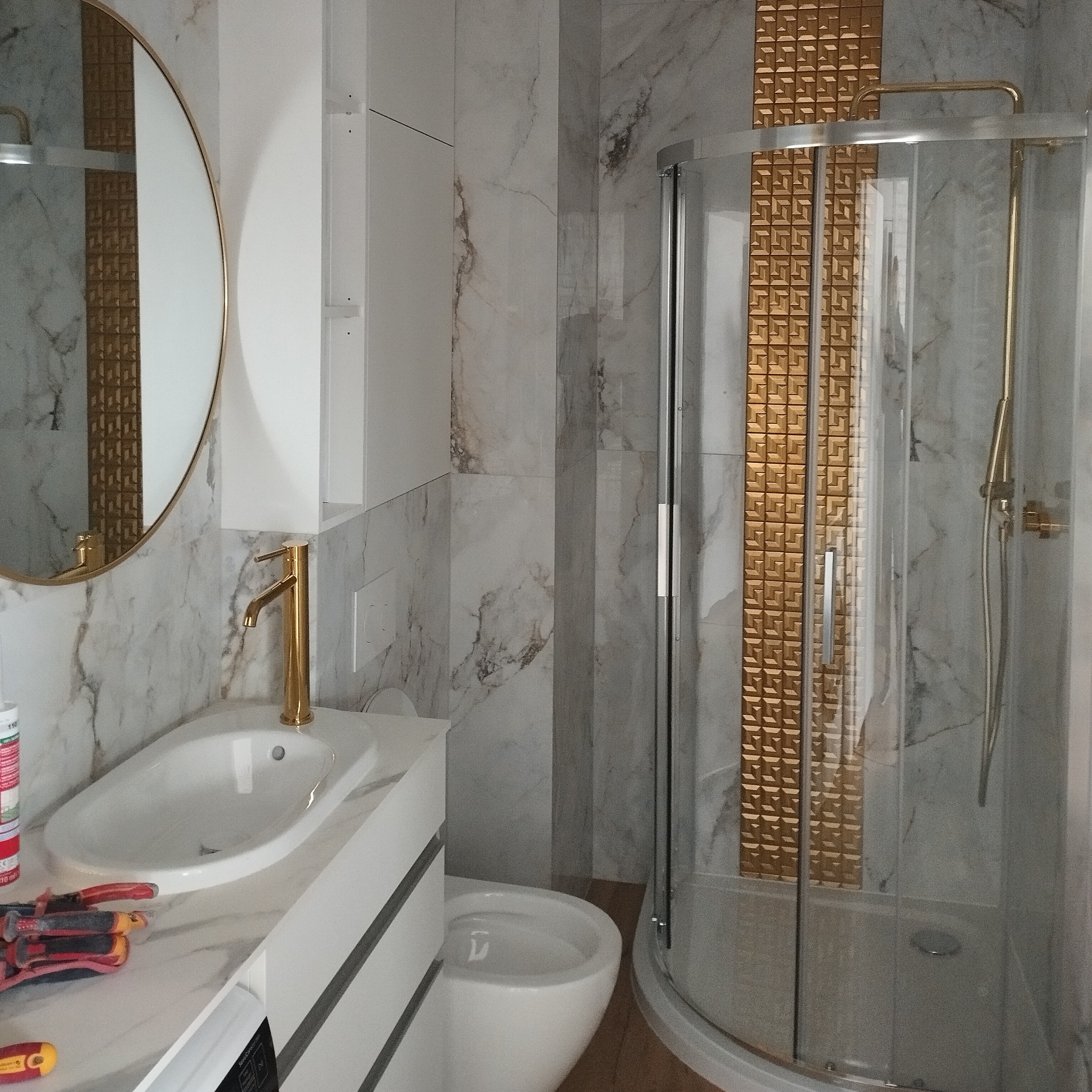 Nowoczesna łazienka z okrągłym lustrem, złotą armaturą, kabiną prysznicową i narzędziami elektrycznymi na blacie umywalki.