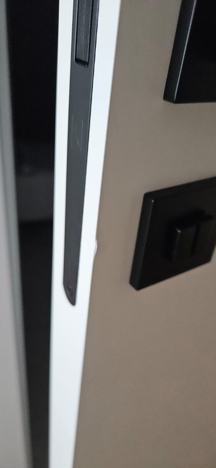 Detal czarnego, kwadratowego włącznika światła na białej ścianie, z widoczną krawędzią drzwi i framugi. Minimalistyczne wykończenie wnętrza.