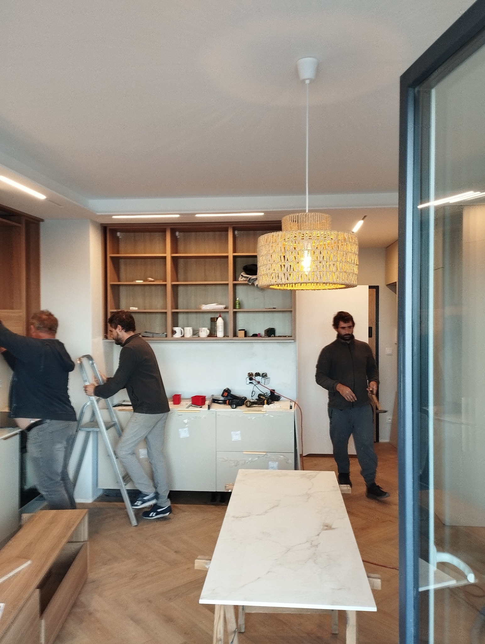 Montaż lampy wiszącej w Krakowie. Dwóch mężczyzn na drabinie, narzędzia na blacie. W tle półki i trzeci mężczyzna. Nowoczesne wnętrze w trakcie wykańczania.