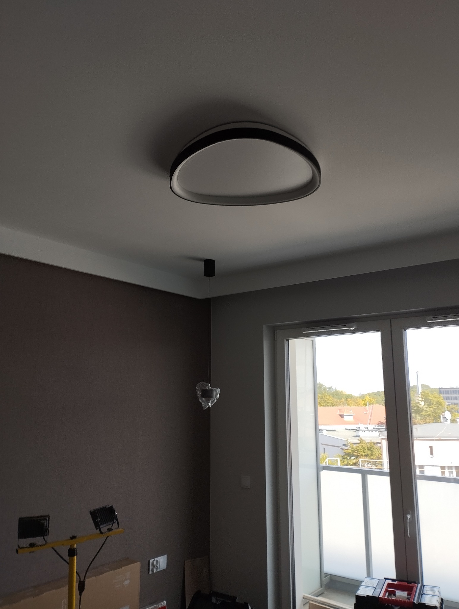 Zamontowana, okrągła lampa sufitowa z czarną obwódką, widoczna w jasnym wnętrzu z oknem balkonowym i narzędziami na podłodze. Minimalistyczny design.