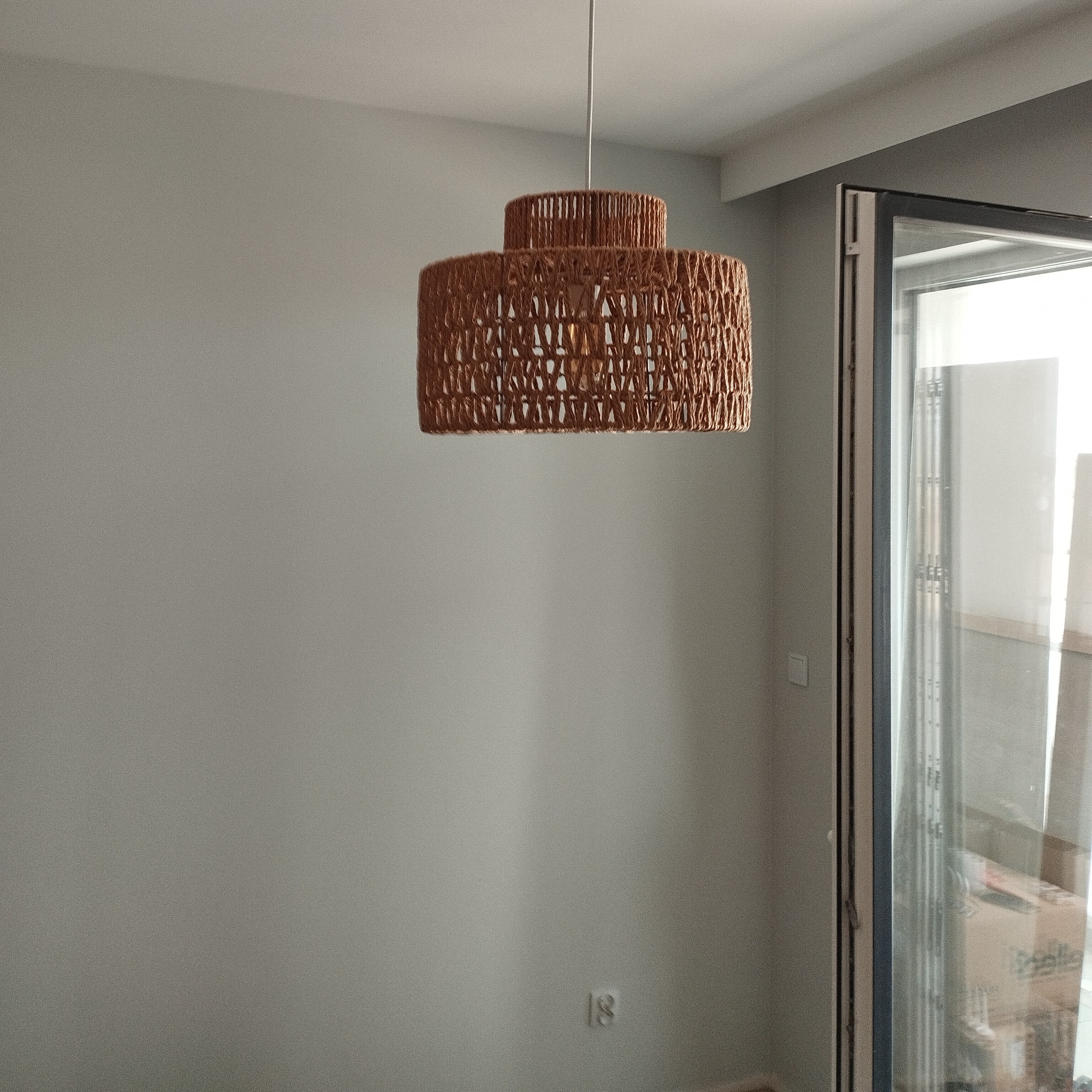 Ręcznie robiona, dwupoziomowa lampa z brązowego sznurka w minimalistycznym wnętrzu z szarą ścianą i oknem balkonowym. Nowoczesne oświetlenie.