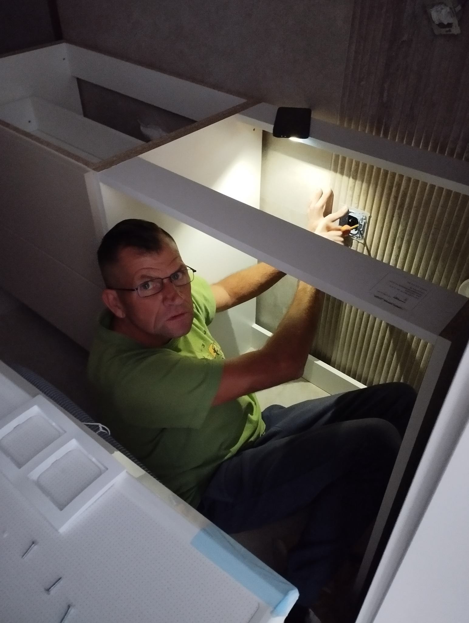 Elektryk w trakcie pracy, montuje gniazdko w szafce podświetlonej lampką. Widoczna ściana z tapetą w tle.