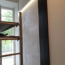 Robert Tomaszek - Ściana z dekoracyjnym tynkiem, oświetleniem LED i pionowymi panelami z czarnego drewna, widoczne rusztowanie malarskie przy oknie.