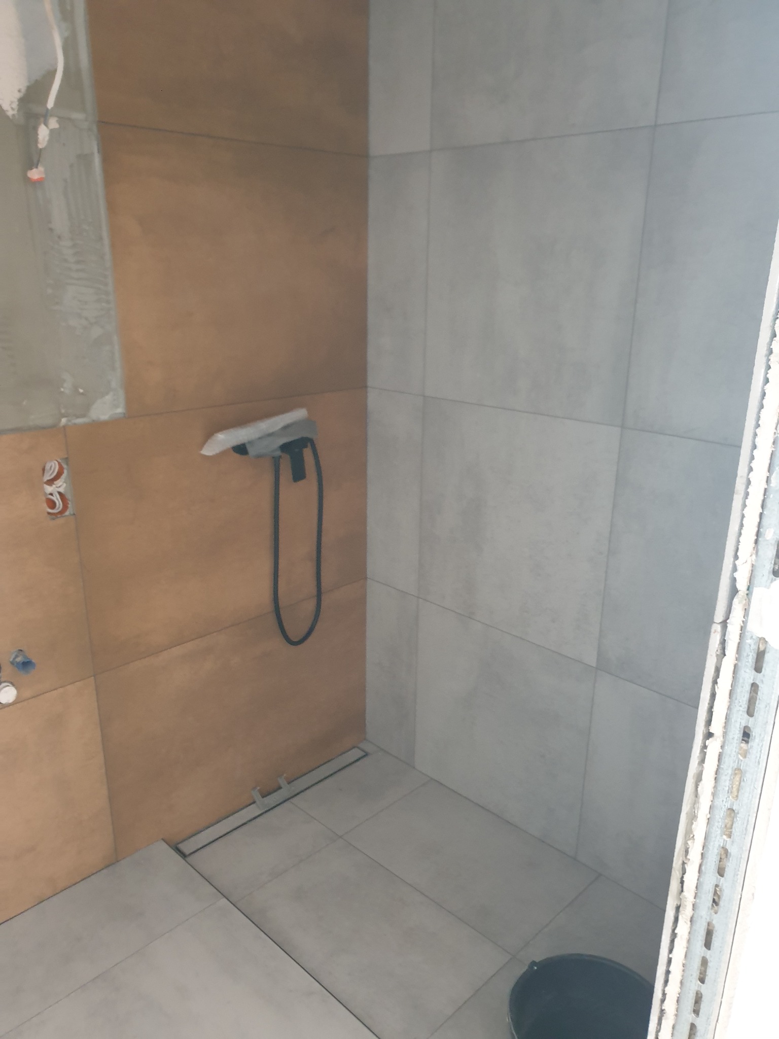 Remontowana łazienka z brązowymi i szarymi płytkami, widoczny odpływ liniowy, niezabezpieczona bateria prysznicowa i wiadro na podłodze.