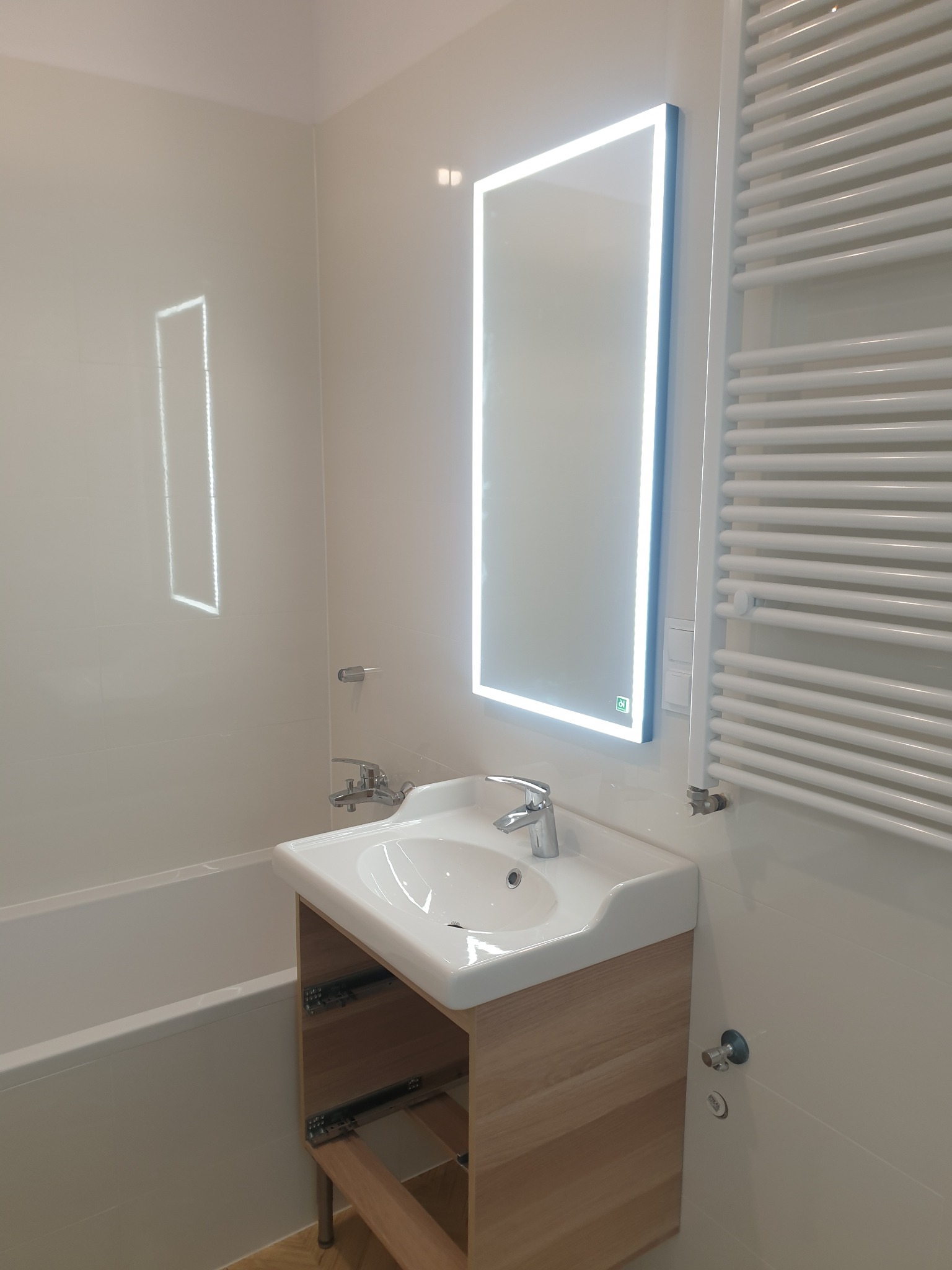 Nowoczesna łazienka z podświetlanym lustrem, białą umywalką na drewnianej szafce, grzejnikiem drabinkowym i wanną obłożoną białymi płytkami.