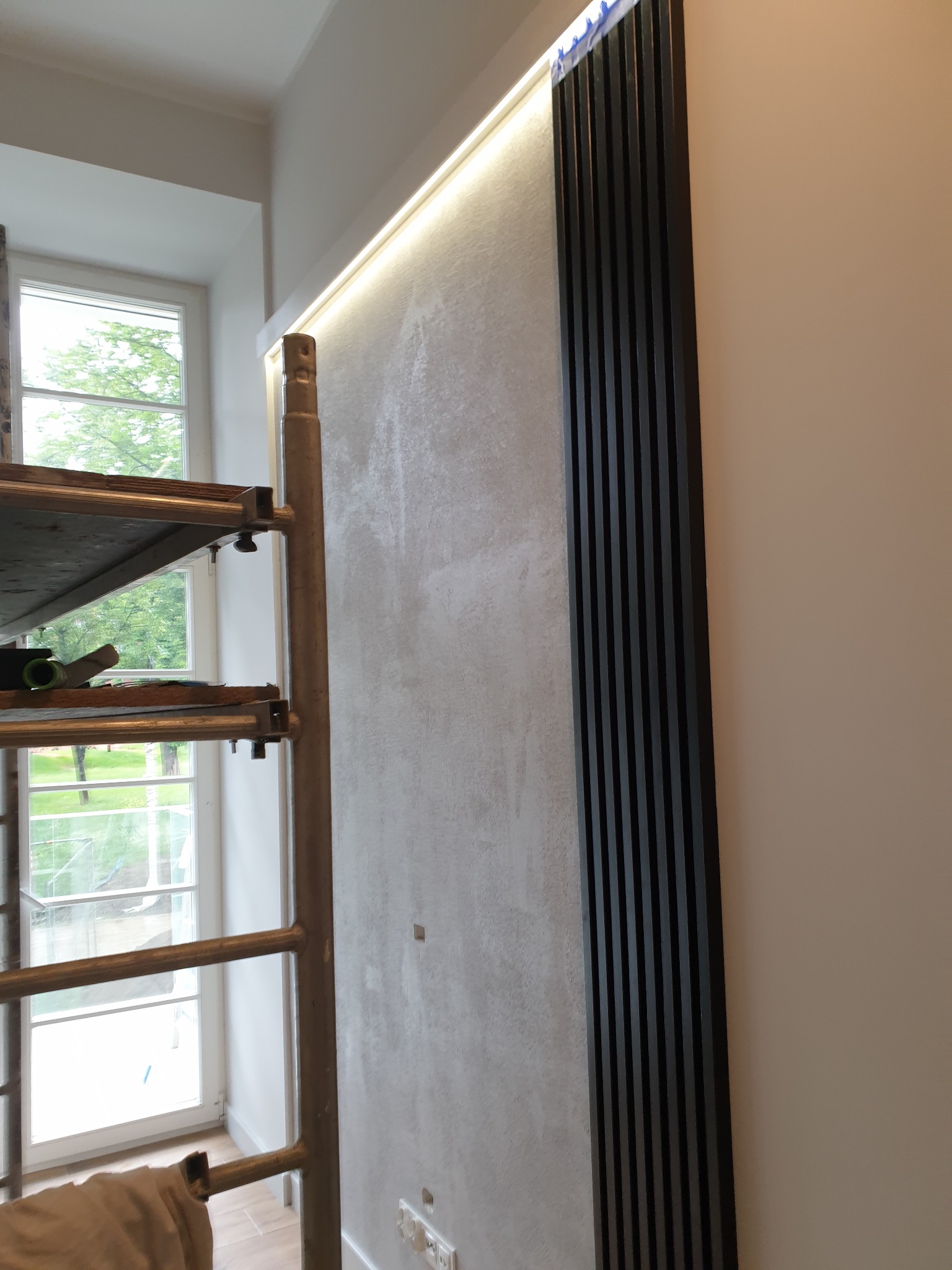 Ściana z dekoracyjnym tynkiem, oświetleniem LED i pionowymi panelami z czarnego drewna, widoczne rusztowanie malarskie przy oknie.