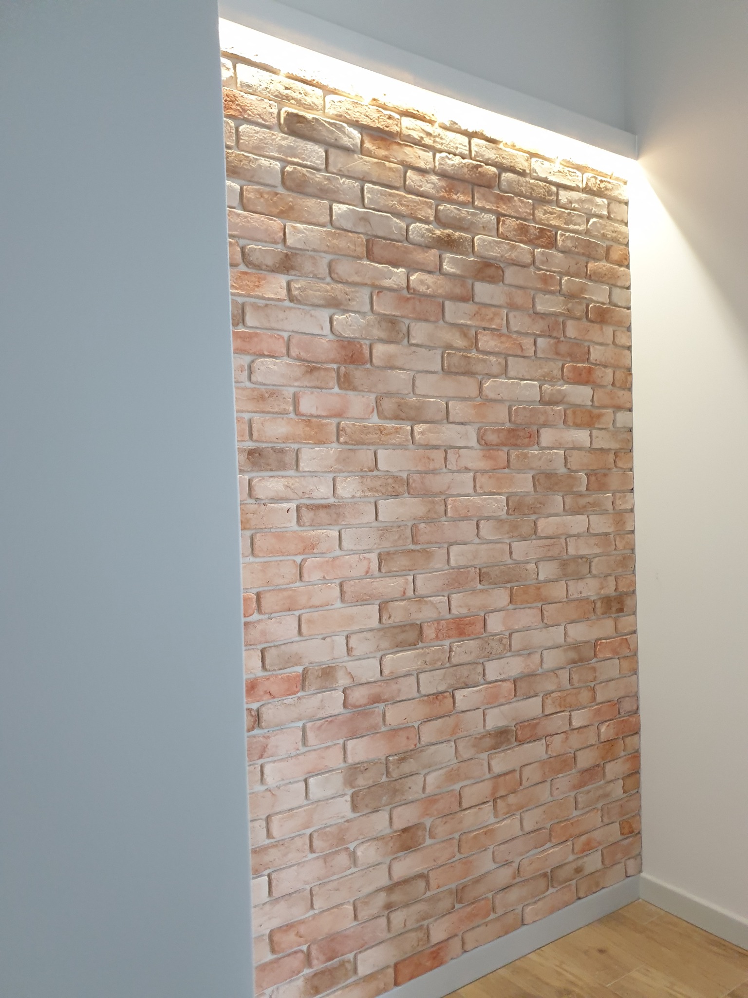 Ściana z cegły klinkierowej w korytarzu z ukrytym oświetleniem LED, widok na narożnik ściany i fragment podłogi z panelami.
