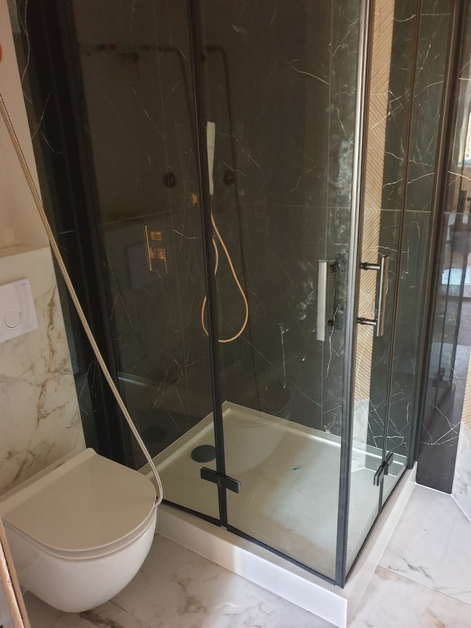 Kabina prysznicowa z czarnymi profilami i marmurowymi płytkami na ścianach, widoczna również toaleta podwieszana z białej ceramiki.