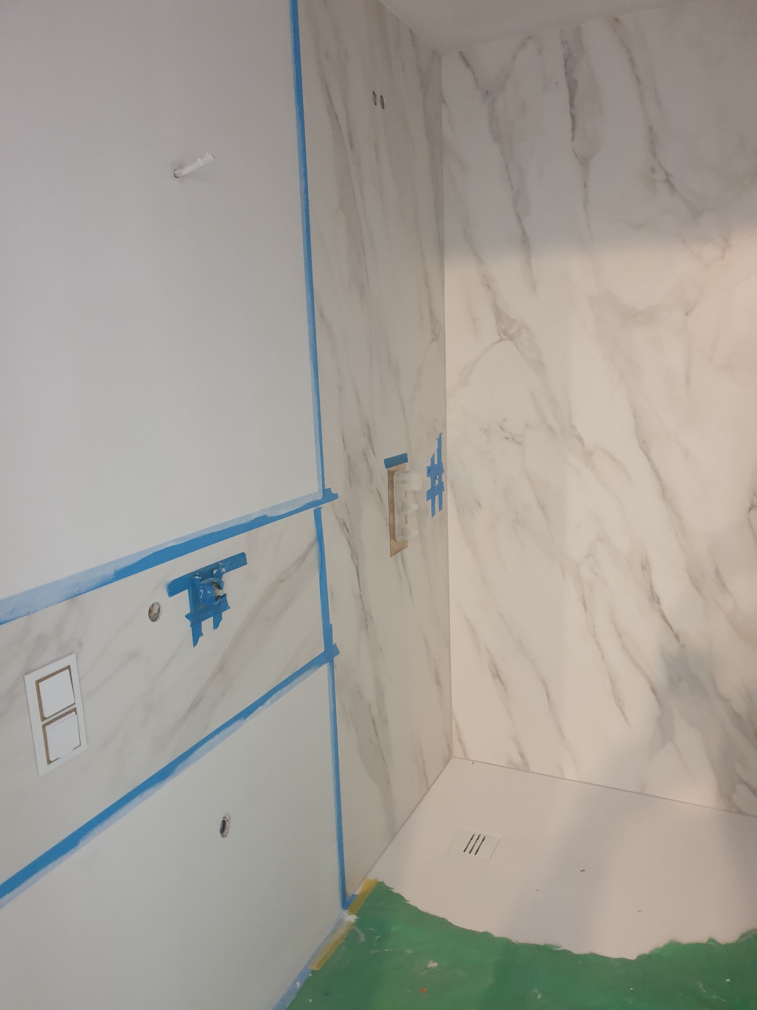 Łazienka w trakcie remontu: ściany wyłożone białymi płytami imitującymi marmur, zabezpieczone niebieską taśmą malarską, widoczny brodzik i instalacja wodna.