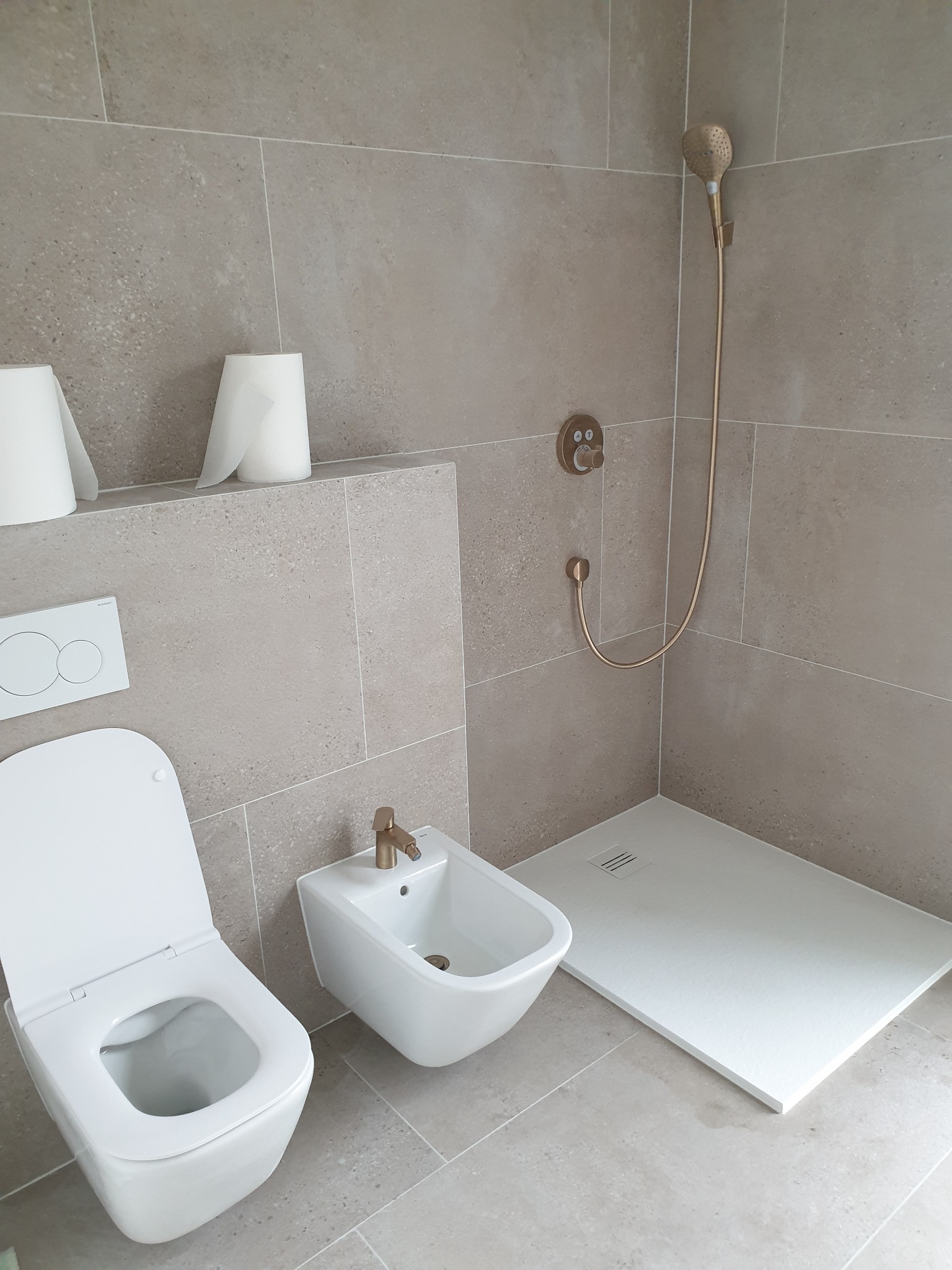 Wykończona łazienka z beżowymi płytkami, wiszącą toaletą z otwartą klapą, bidetem z mosiężną armaturą, prysznicem typu walk-in z mosiężną słuchawką i termostatem oraz dwoma rolkami papieru...