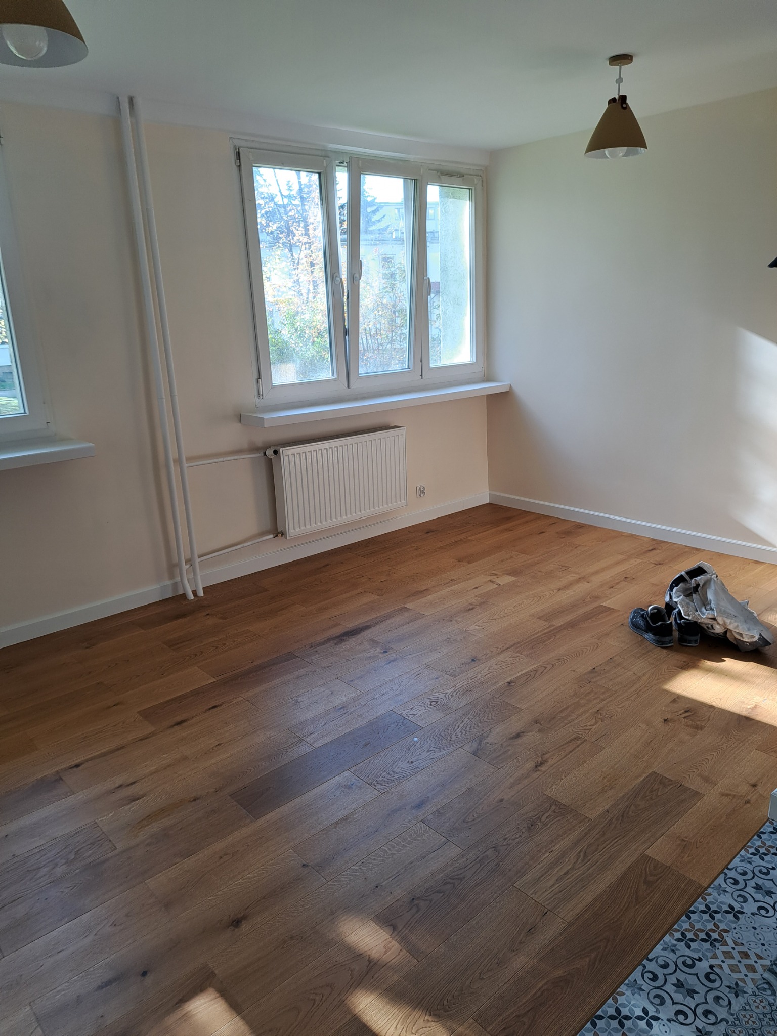 Pokój po remoncie z nową, drewnianą podłogą; widoczne okno z parapetem, grzejnik, lampa sufitowa w stylu skandynawskim oraz ubrania robocze i buty leżące na podłodze.