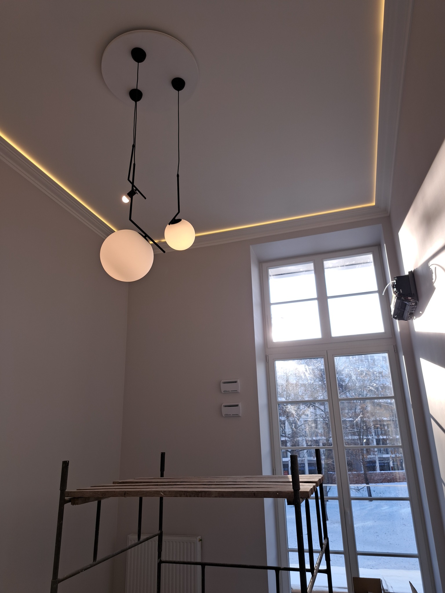 Wnętrze w trakcie prac: widok na sufit z dekoracyjnym oświetleniem LED wzdłuż krawędzi, nowoczesna lampa wisząca z dwoma kulistymi kloszami, rusztowanie budowlane w dolnej części kadru i duże okno...