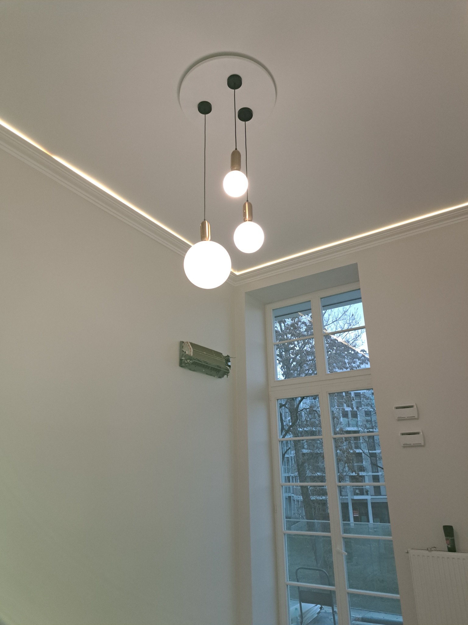 Jasne wnętrze z białymi ścianami, oświetleniem LED wzdłuż sufitu i trzema wiszącymi lampami kulami. Widoczne ślady po demontażu klimatyzacji na ścianie i okno z widokiem na budynek.