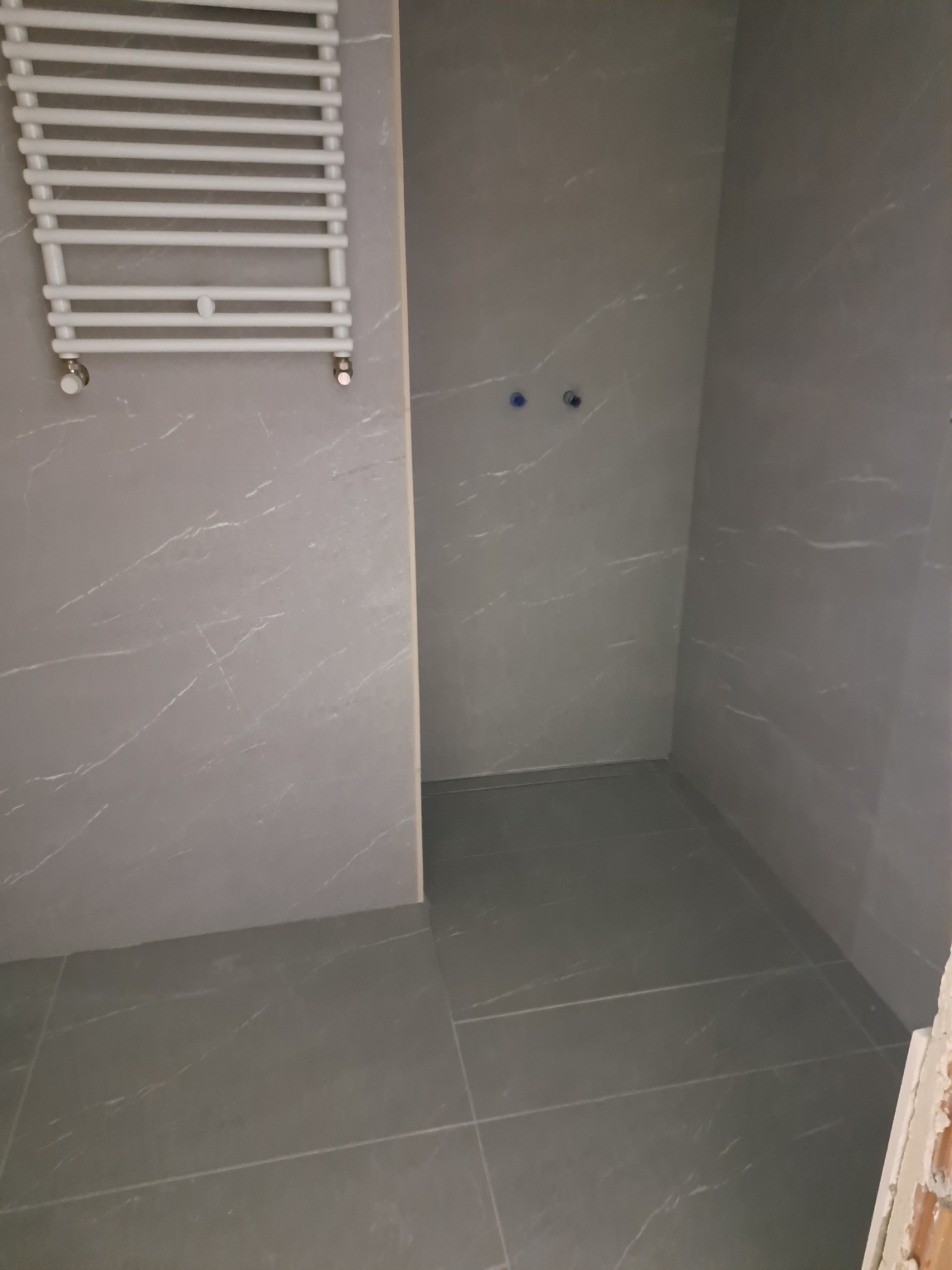 Wykończona łazienka z szarymi płytkami imitującymi marmur, widoczny grzejnik drabinkowy i przygotowane punkty instalacji wodnej.