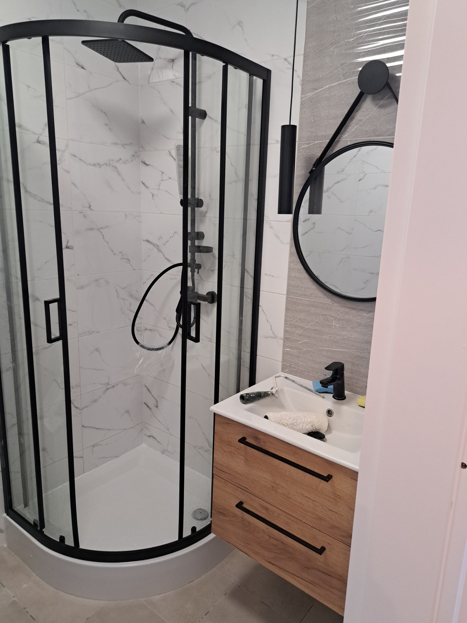 Nowoczesna łazienka po remoncie: kabina prysznicowa z czarnymi profilami, umywalka z drewnianą szafką i czarną armaturą, okrągłe lustro na ścianie z marmurowym wzorem, akcesoria malarskie w umywalce.
