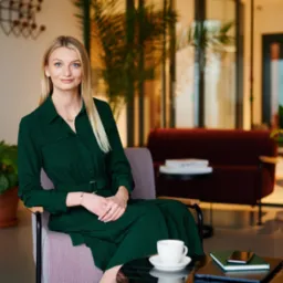 Profesjonalna blondynka w zielonej sukience siedzi w fotelu, z filiżanką kawy i notatnikiem na stoliku, w tle nowoczesne biuro z roślinami doniczkowymi.