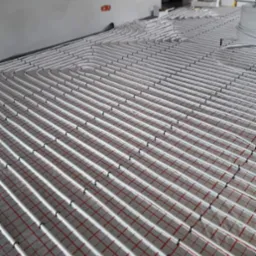 Rozłożone rury ogrzewania podłogowego na izolacji z siatką w nowym budynku, widoczne mocowania rur do podłoża, fragment ściany i przygotowania do wylewki.