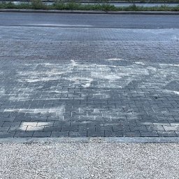 SPEC BUD Patryk Gołuch - Obszar z szarej kostki brukowej, częściowo pokrytej białym nalotem, z widocznym małym stosem kostki na palecie i osobą w odblaskowej kamizelce w tle. Krawędź wyłożona drobnym żwirem.