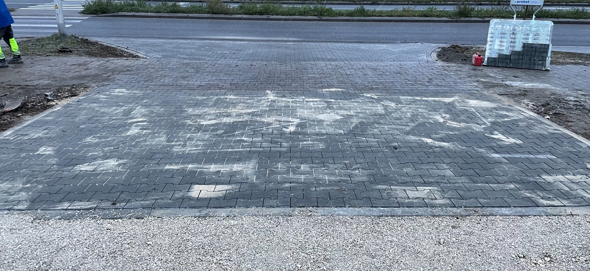 Obszar z szarej kostki brukowej, częściowo pokrytej białym nalotem, z widocznym małym stosem kostki na palecie i osobą w odblaskowej kamizelce w tle. Krawędź wyłożona drobnym żwirem.
