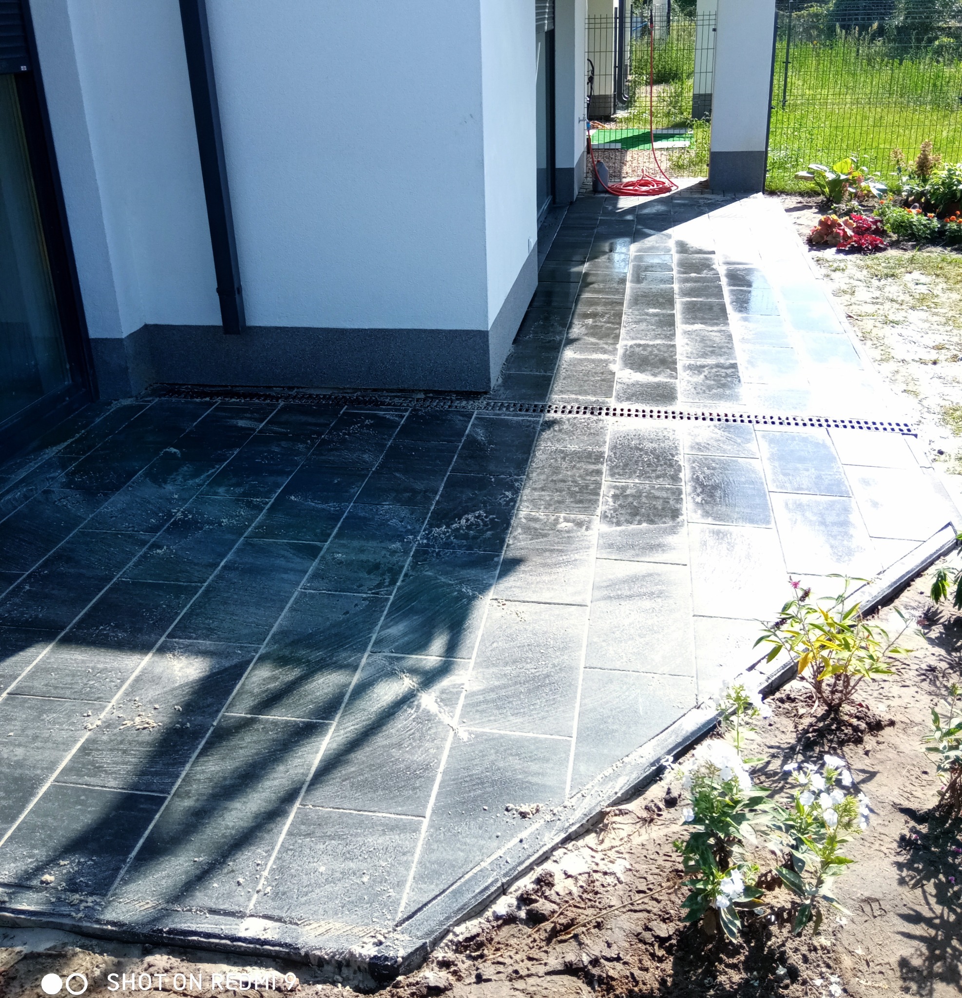 Nowo ułożona, grafitowa kostka brukowa na tarasie przydomowym z odpływem liniowym, widoczne świeżo posadzone kwiaty i krzewy.