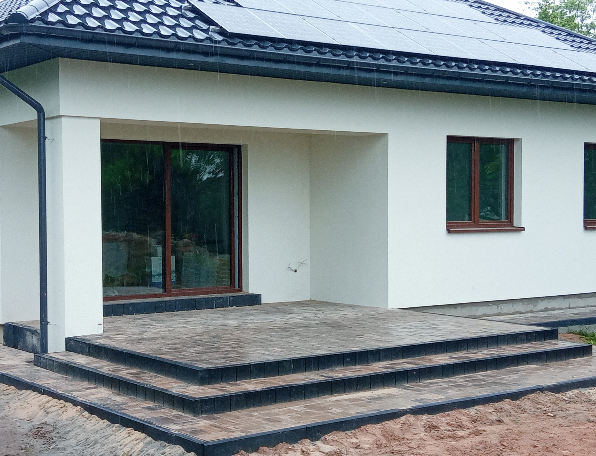 Nowoczesny dom z białymi ścianami i schodami wejściowymi wyłożonymi kostką brukową w odcieniach szarości i czerni, widok w pochmurny deszczowy dzień.