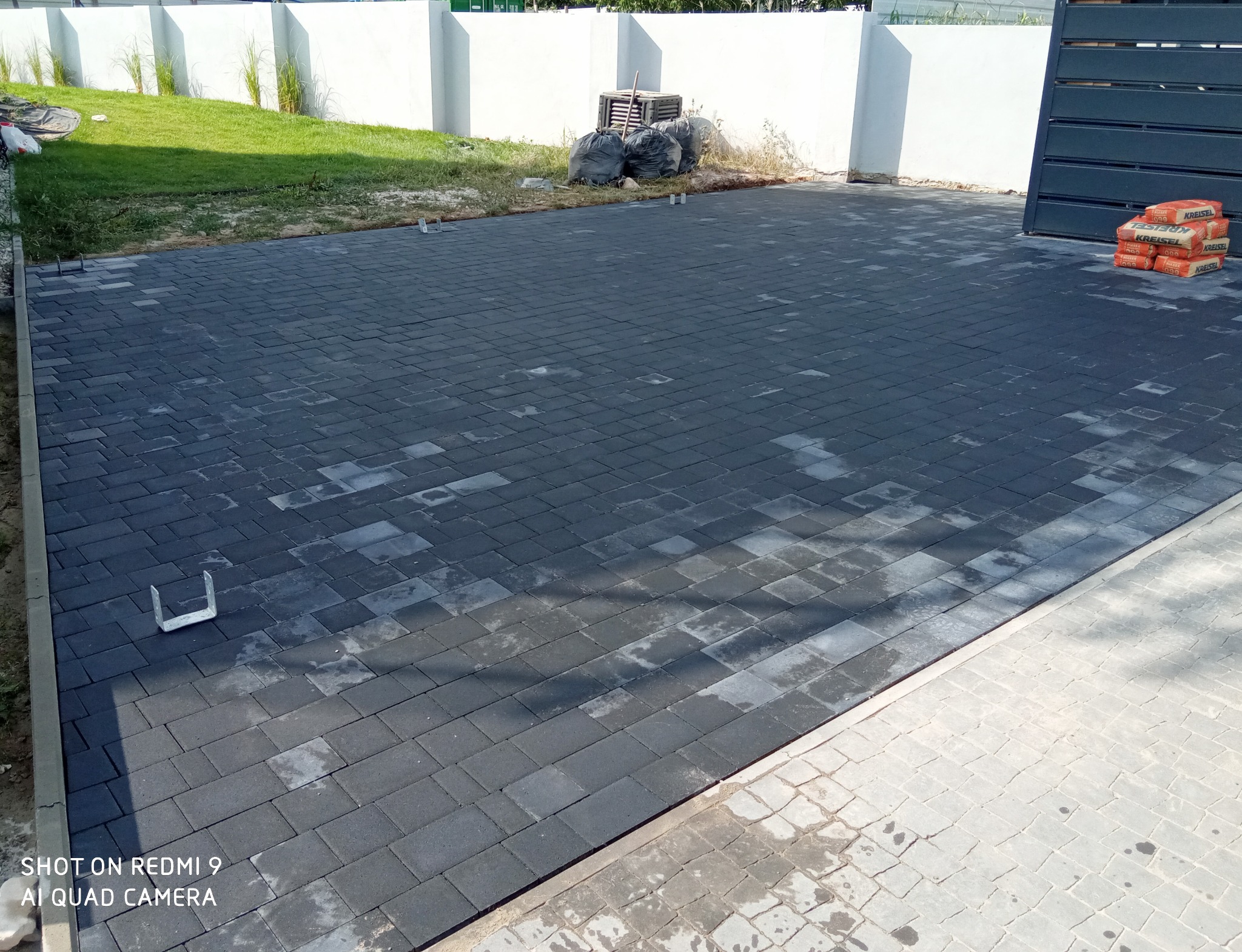 Częściowo ułożona, ciemnoszara kostka brukowa na podjeździe, widoczne worki z cementem i narzędzia montażowe, trawa i biały mur w tle.
