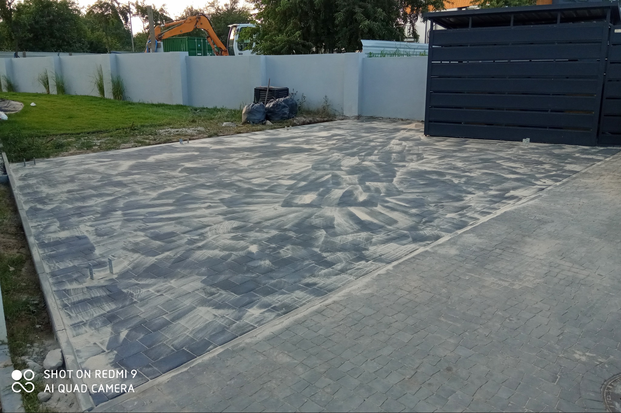 Świeżo ułożona, szara kostka brukowa z widocznymi śladami po piasku do wypełniania szczelin, na tle szarego ogrodzenia i bramy wjazdowej, z koparką w oddali.