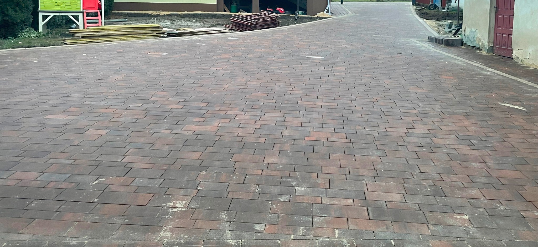 Ułożona, brązowo-czerwona kostka brukowa na podjeździe, widoczne przygotowane deski i elementy placu zabaw w tle.