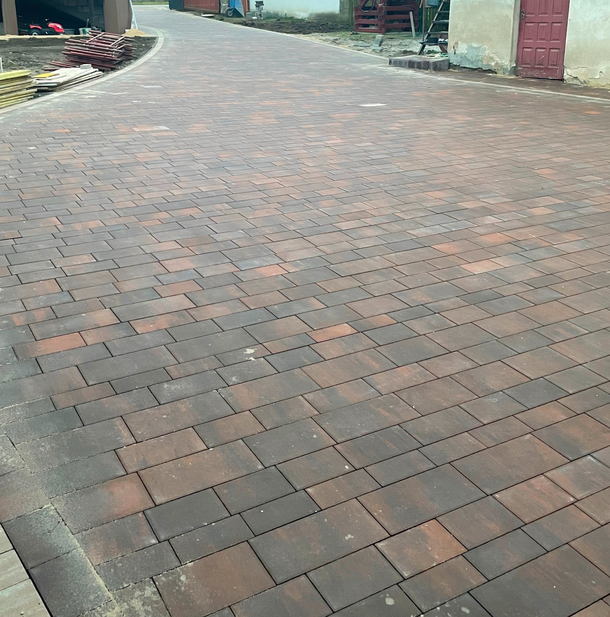 Szeroki, zakrzywiony podjazd wyłożony brązowo-szarą kostką brukową w kształcie prostokątów, z widocznymi elementami przygotowanymi do dalszych prac po bokach, w tle budynki.