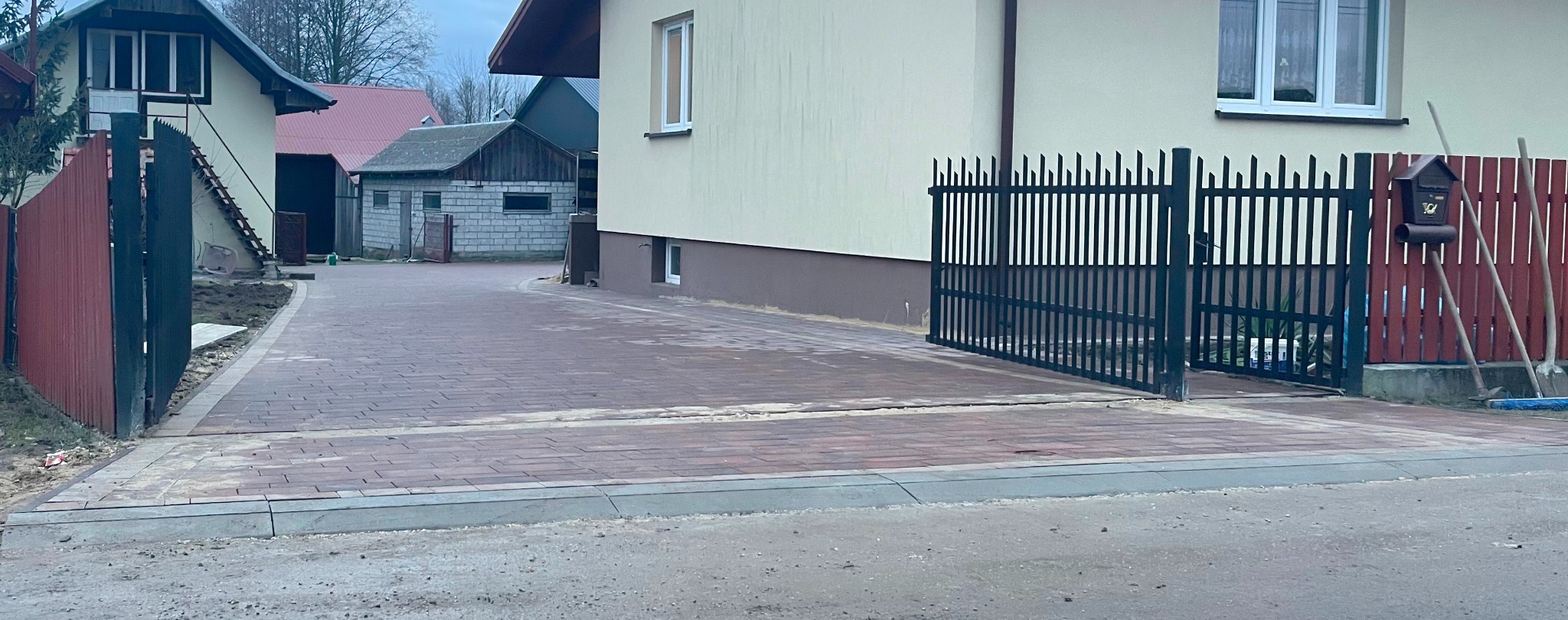 Ułożona, czerwono-brązowa kostka brukowa na podjeździe posesji, z widocznymi obrzeżami betonowymi, metalową bramą wjazdową i fragmentami ogrodzenia oraz budynków w tle.