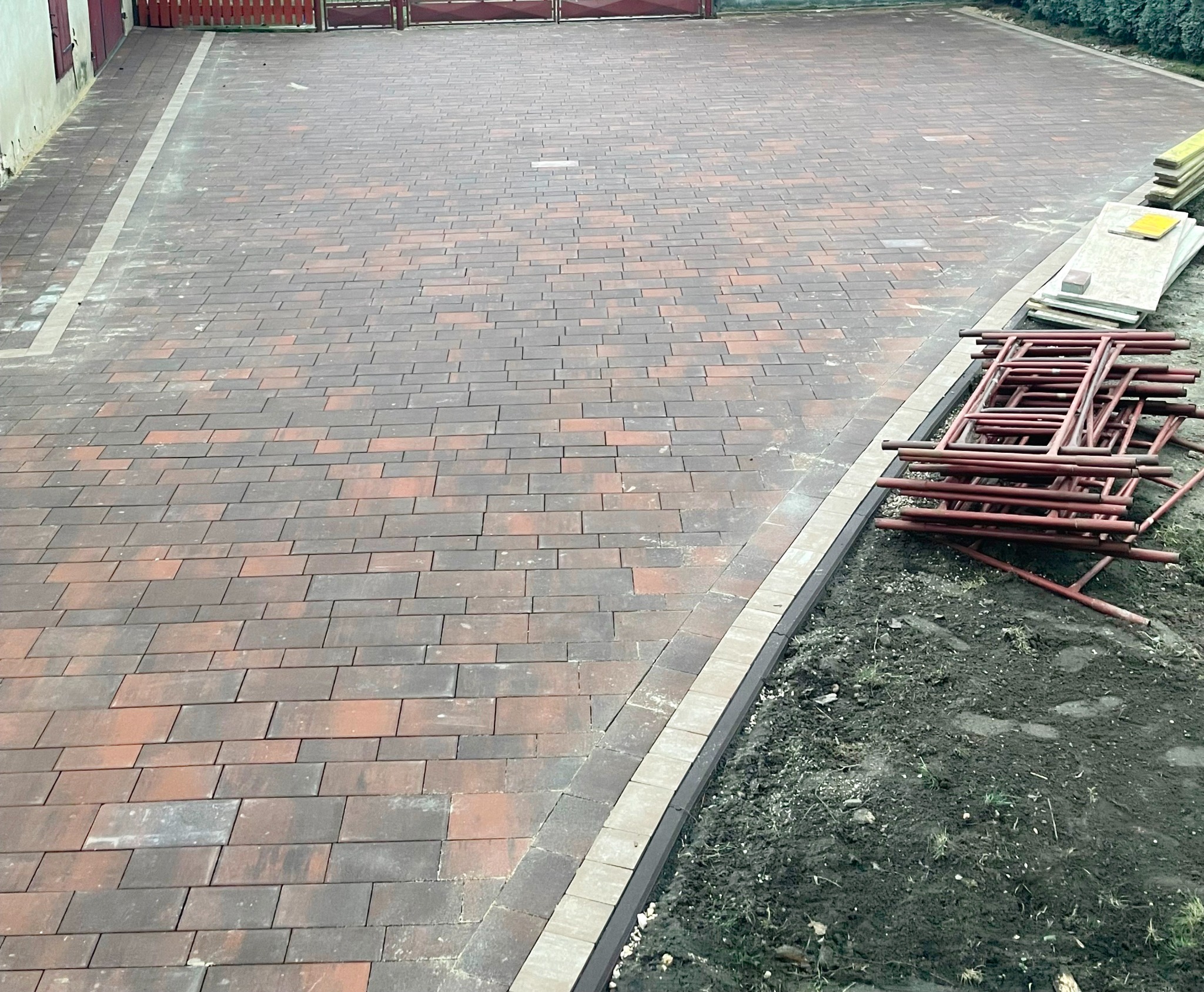Nowo ułożona, dwukolorowa kostka brukowa na posesji, widoczne obrzeża i przygotowane podłoże z boku, na którym leżą metalowe podpory.